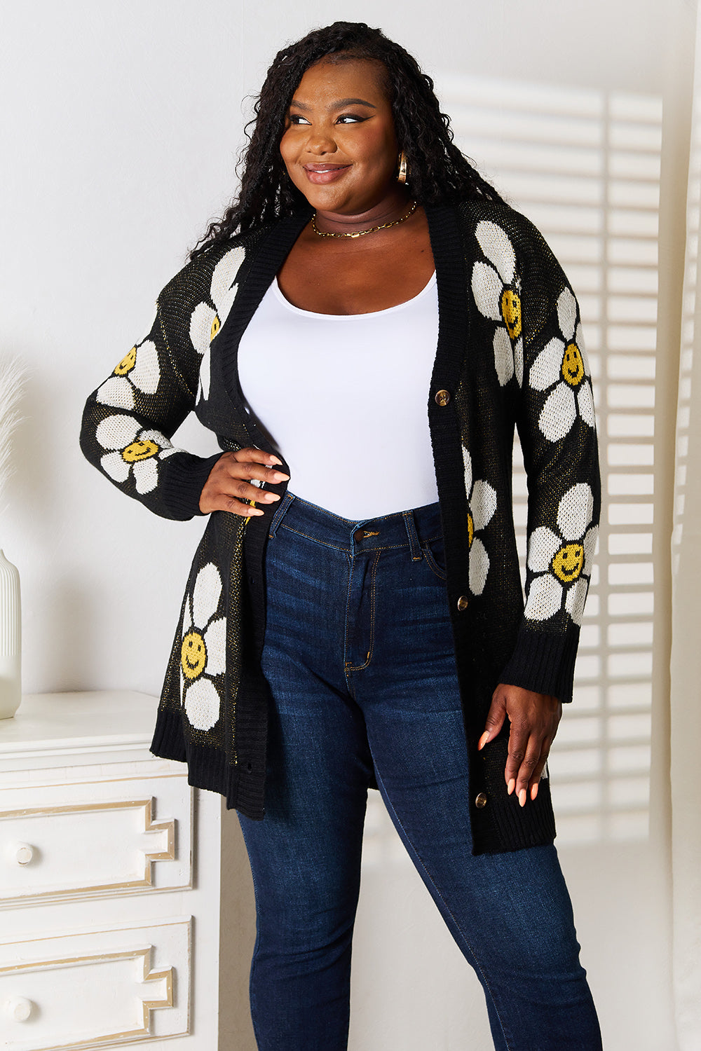 A woman stands indoors wearing the Perfee Floral Button Down Longline Cardigan, which features stylish black fabric adorned with white and yellow smiley face flowers. She pairs it with a white top and blue jeans, looking off to the side while smiling.