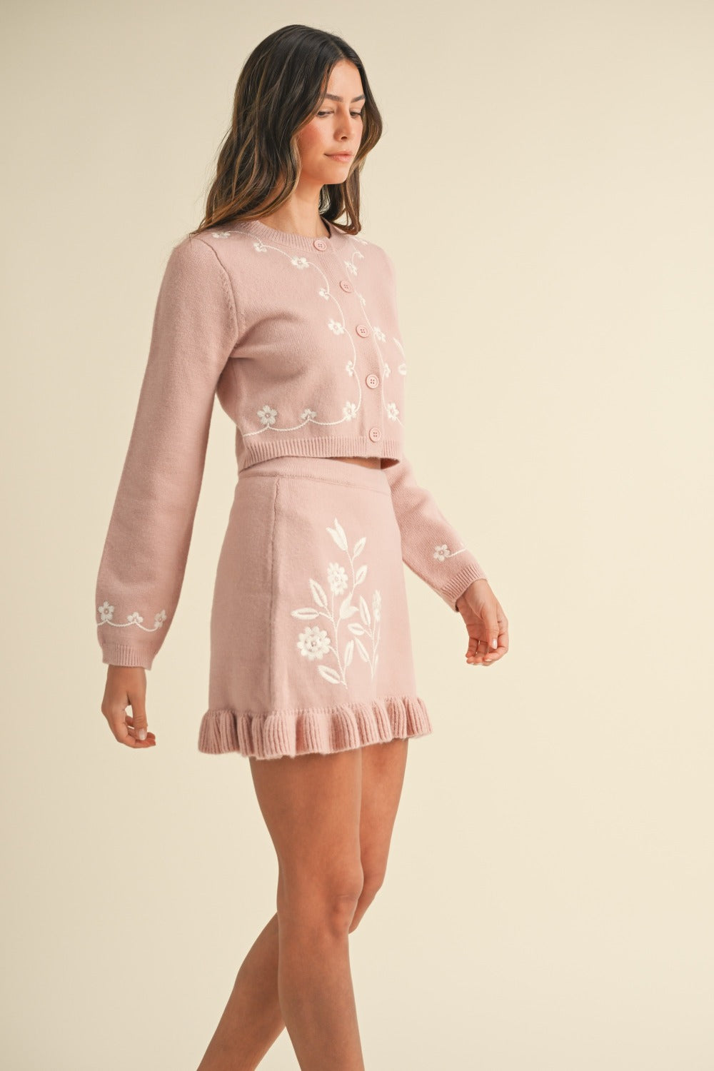 A woman is wearing the MABLE Floral Embroidered Crop Cardigan and Ruffled Mini Skirt Set, showcasing a pink two-piece ensemble with detailed embroidered flower patterns. She has long hair and is posing against a plain background.