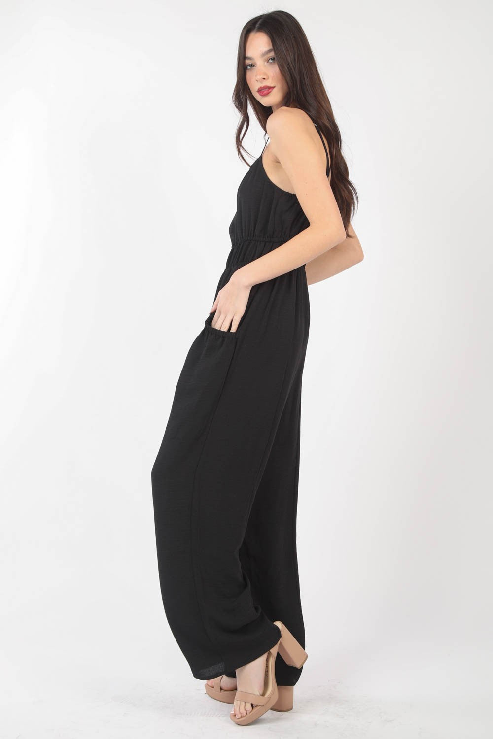 A woman stands against a plain backdrop wearing the VERY J Pintuck Detail Woven Sleeveless Jumpsuit. The black jumpsuit has thin straps, wide-leg pants, and is crafted from woven fabric with subtle pintuck details. She has her hands in her pockets and is looking at the camera.