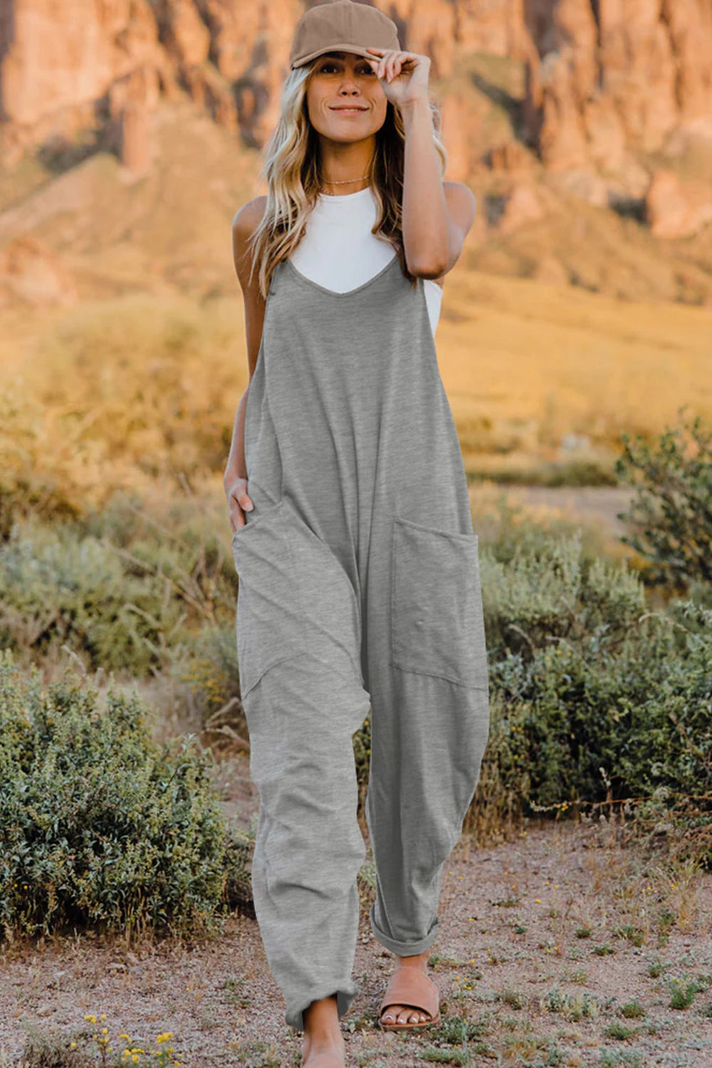 Person wearing a Double Take Full Size V-Neck Sleeveless Jumpsuit with Pockets and sandals, walking outdoors on a dirt trail with bushes in the background.