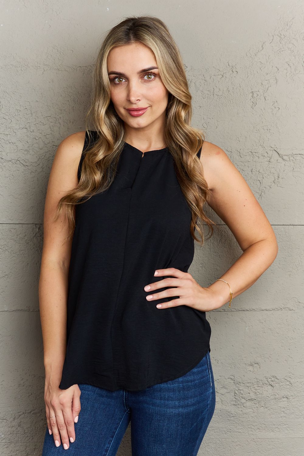 A woman with long blonde hair stands against a textured wall, wearing the Ninexis First Glance Sleeveless Neckline Slit Top in black and blue jeans. Her left hand is on her hip while her right hand rests on the wall.