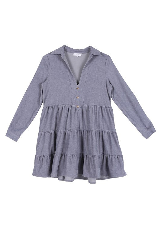 The woman in the Corduroy tiered dress, featuring a deep V-neck, smiles as she gently touches her forehead, showcasing the light blue fabric cascading elegantly.