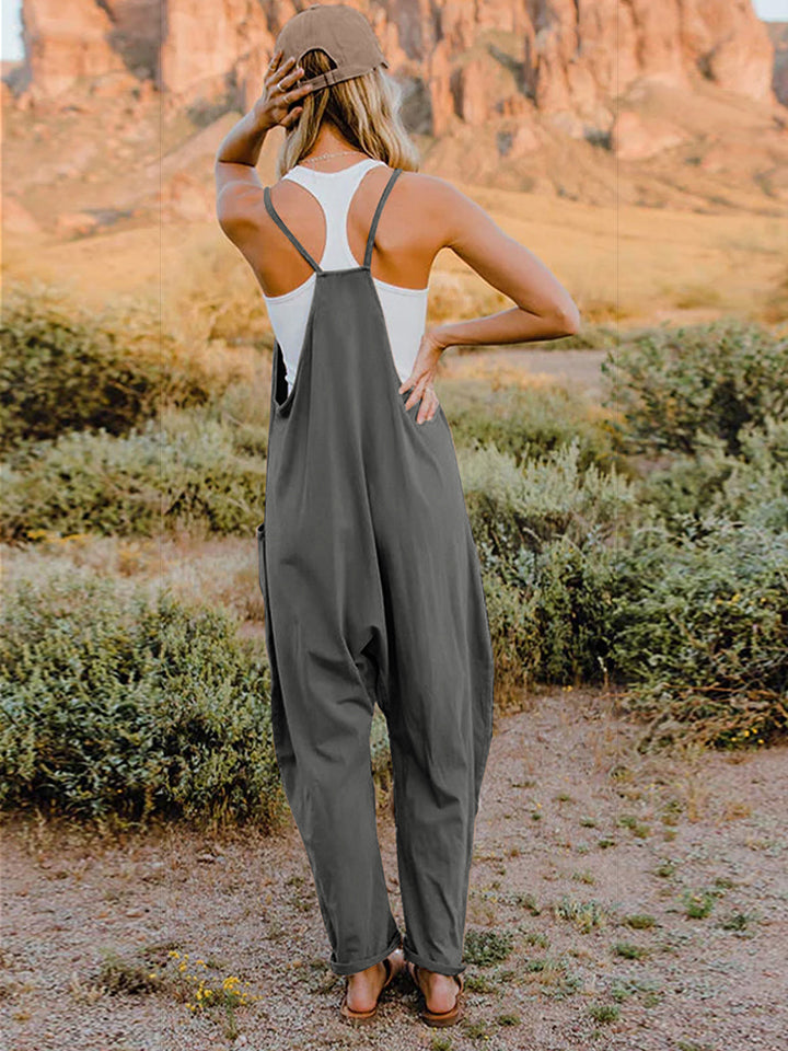 A woman wearing the Double Take Full Size Sleeveless V-Neck Pocketed Jumpsuit in green and a beige cap stands outdoors in a desert landscape with plants and rocky cliffs in the background.