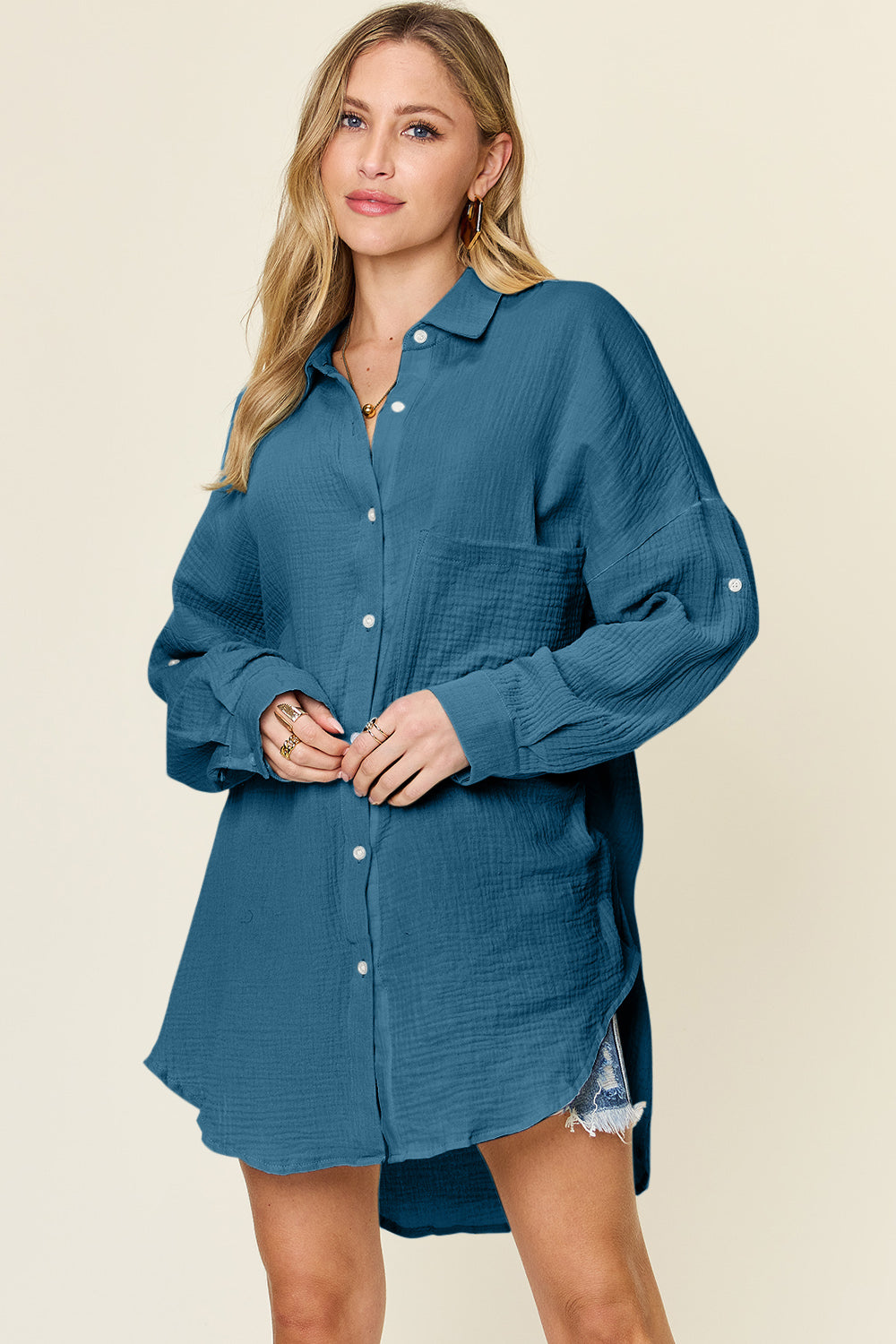 A person is wearing the Double Take Full Size Pocketed Texture Button Up Shirt, a long-sleeved, color-blocked piece made of 100% cotton in shades of green and cream. They have paired it with distressed denim shorts and are standing against a neutral backdrop. The shirt features buttoned cuffs and offers a relaxed yet stylish look. It is easy to care for with machine wash cold instructions.