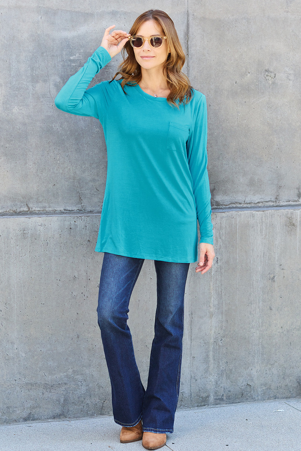 A woman with long hair wearing sunglasses, a Basic Bae Full Size Round Neck Long Sleeve Top in turquoise, and jeans is standing in front of a concrete wall with one hand on her hip.