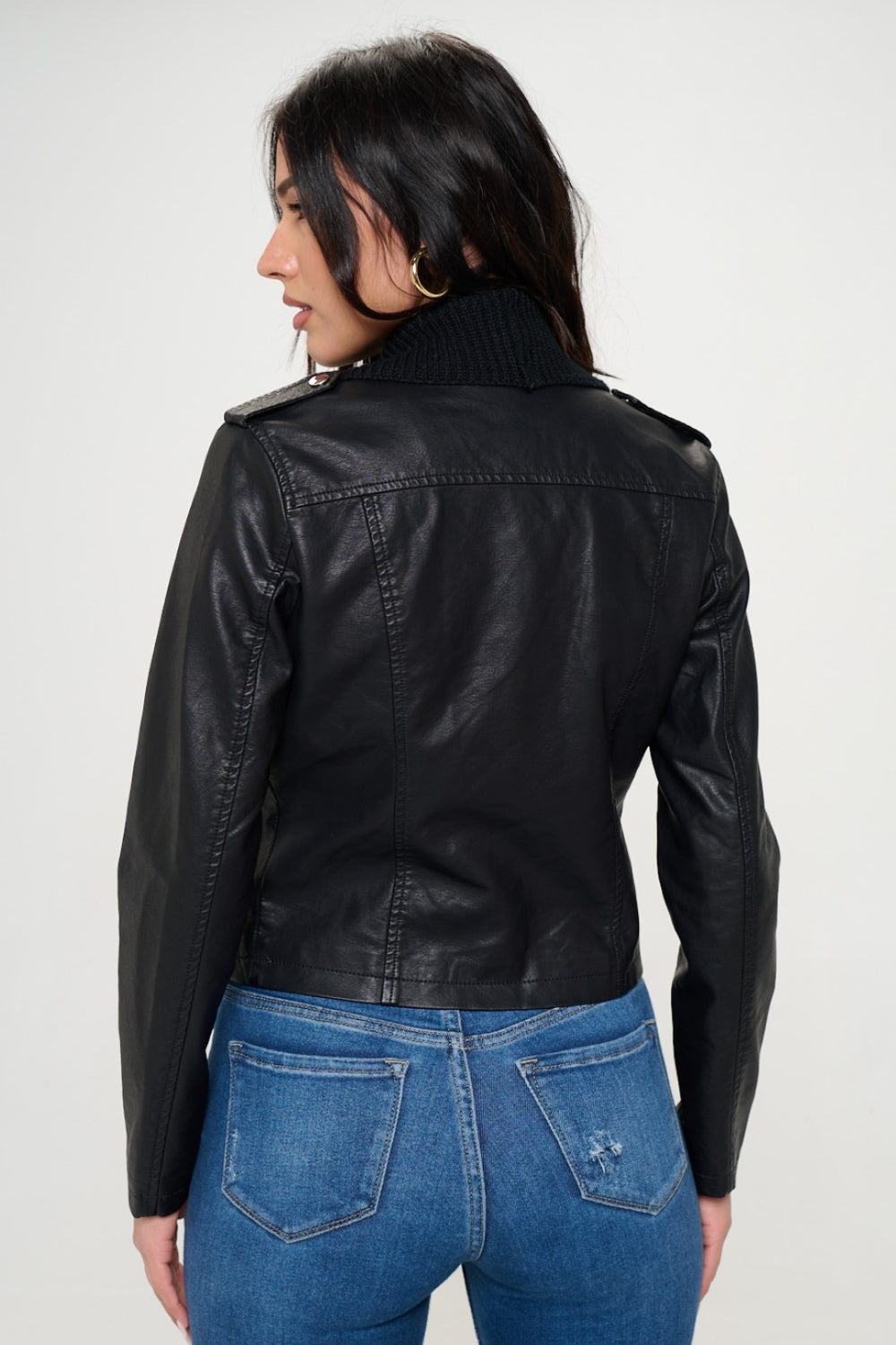 A woman with long, dark hair wearing a black top, a black shawl over the Coalition LA Knit Collared Faux Leather Crop Jacket, and blue jeans stands against a plain white background.