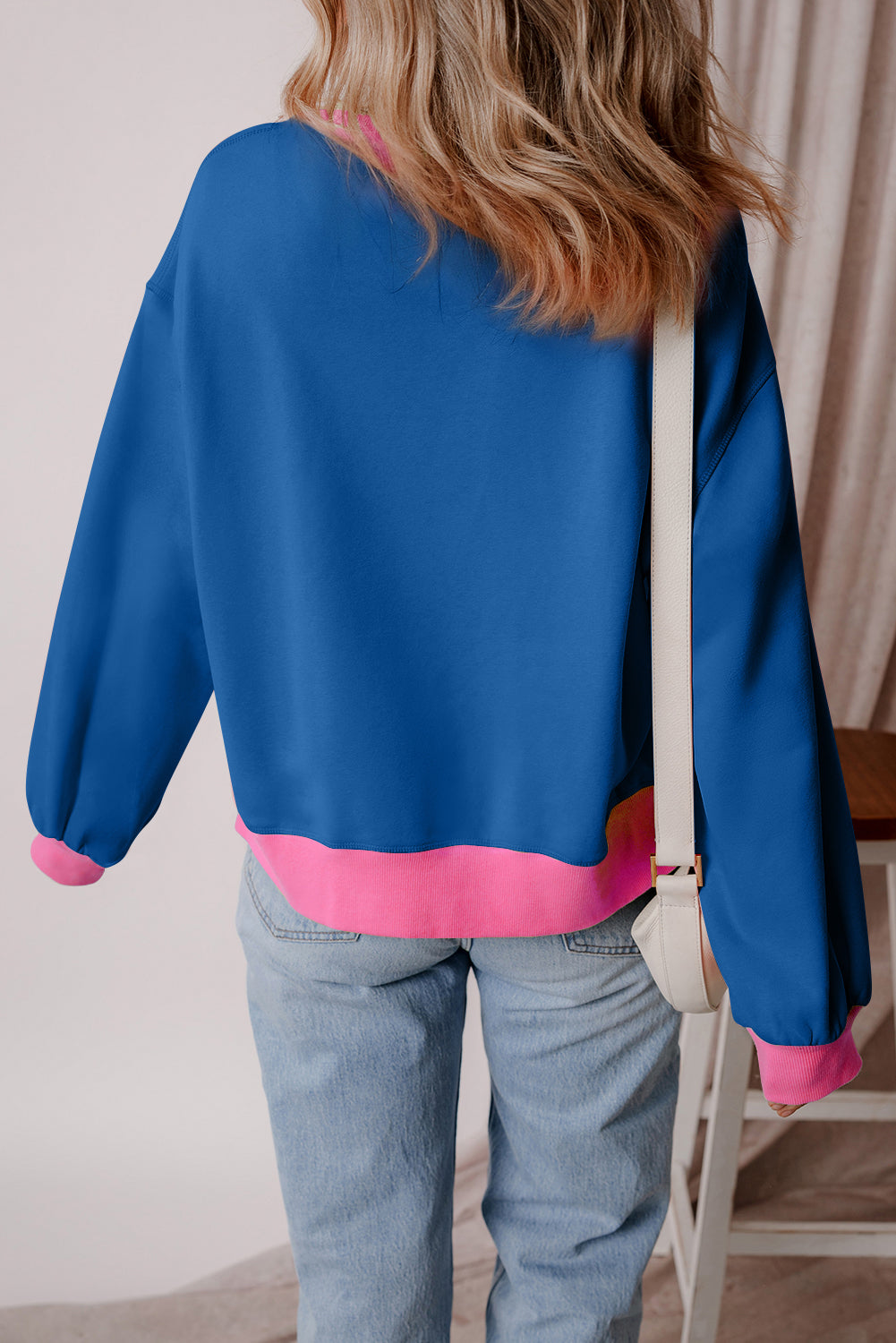 A woman with long blonde hair is smiling as she poses against a purple background, wearing the Blue Colorblock Bubble Sleeve Sweatshirt paired with white pants. The sweatshirt features a boxy fit with pink accents and is ideal for the autumn and winter seasons.