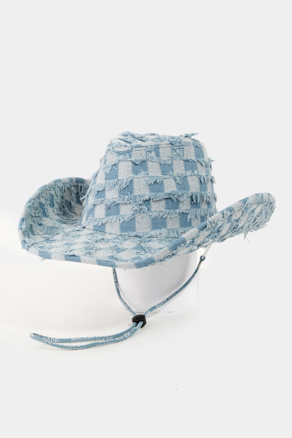 The Fame Checkered Fringe Denim Cowboy Hat, featuring a wide brim and adorned with a blue checkered pattern along with frayed denim edges, rests on a pristine white surface. It includes an adjustable inner string and a matching blue chin strap for added style and functionality.