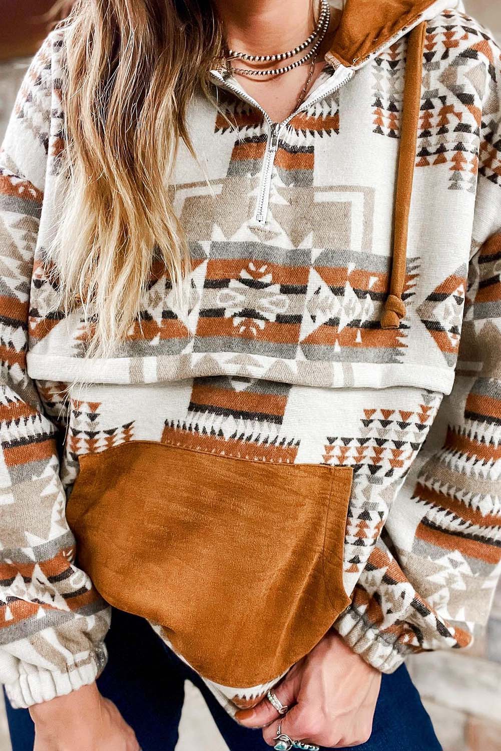 A person with long blonde hair, seen from behind, is wearing the Beige Aztec Print Kangaroo Pocket Half-Zip Hoodie. The hoodie showcases an Aztec pattern in shades of brown, beige, and orange and features a drop shoulder design with a cozy kangaroo pocket, all while standing outdoors.