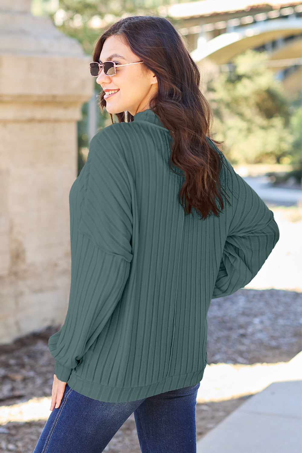 A woman with long hair is wearing a green Basic Bae Full Size Ribbed Exposed Seam Mock Neck Knit Top, blue jeans, and sunglasses. She is standing outdoors and smiling.