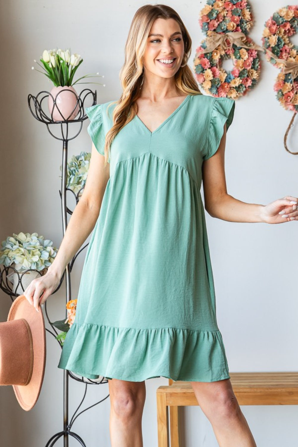 A person in a Heimish Full Size Short Sleeve V Neck Ruffled Hem Dress stands indoors, elegantly holding a hat. The floral decor in the background complements the dress's feminine charm.