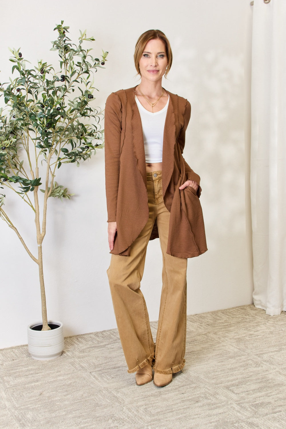 A woman stands in front of a light-colored wall and a small green plant, wearing the Culture Code Full Size Open Front Long Sleeve Cardigan with a merrow edge finish over a white top and beige pants.