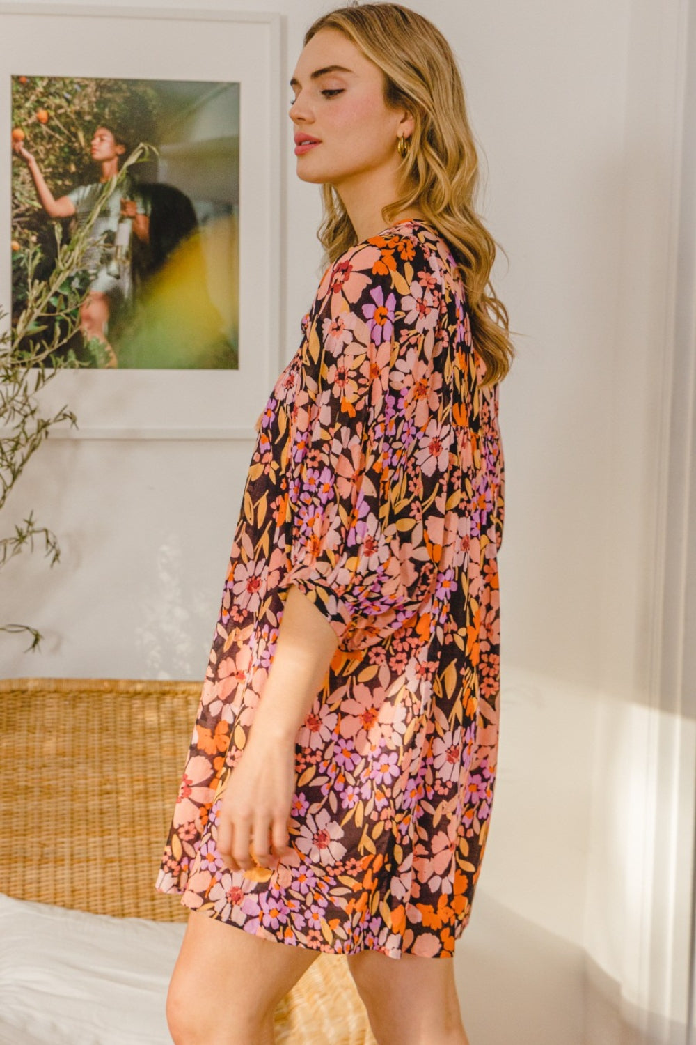 A woman in a flattering silhouette stands indoors with arms folded, wearing the vibrant ODDI Full Size Floral Tied Neck Mini Dress. She poses gracefully in front of a framed picture and wicker chair.