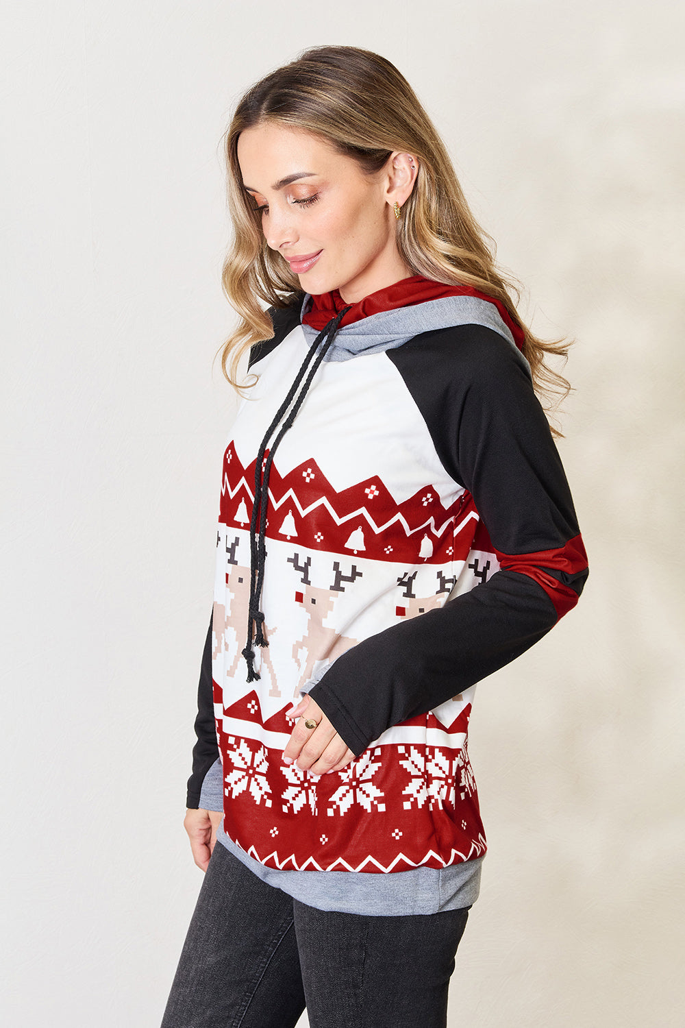 A person is wearing the Double Take Full Size Christmas Drawstring Long Sleeve Hoodie, which features a holiday-themed red and white pattern with reindeer and snowflakes. This cozy hoodie, made from a polyester spandex blend, has stylish black sleeves accented with red stripes. The person is smiling and looking to the side.