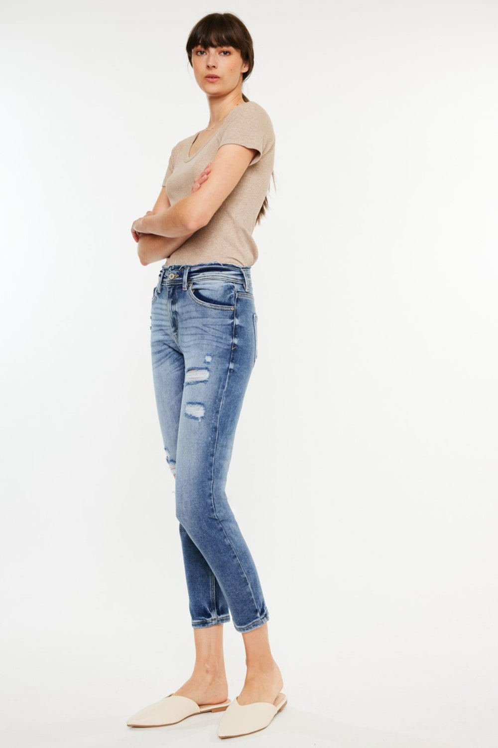 A person stands in front of a plain background, wearing a striped shirt over a beige top, Kancan High Rise Distressed Mom Jeans, and white slip-on shoes.