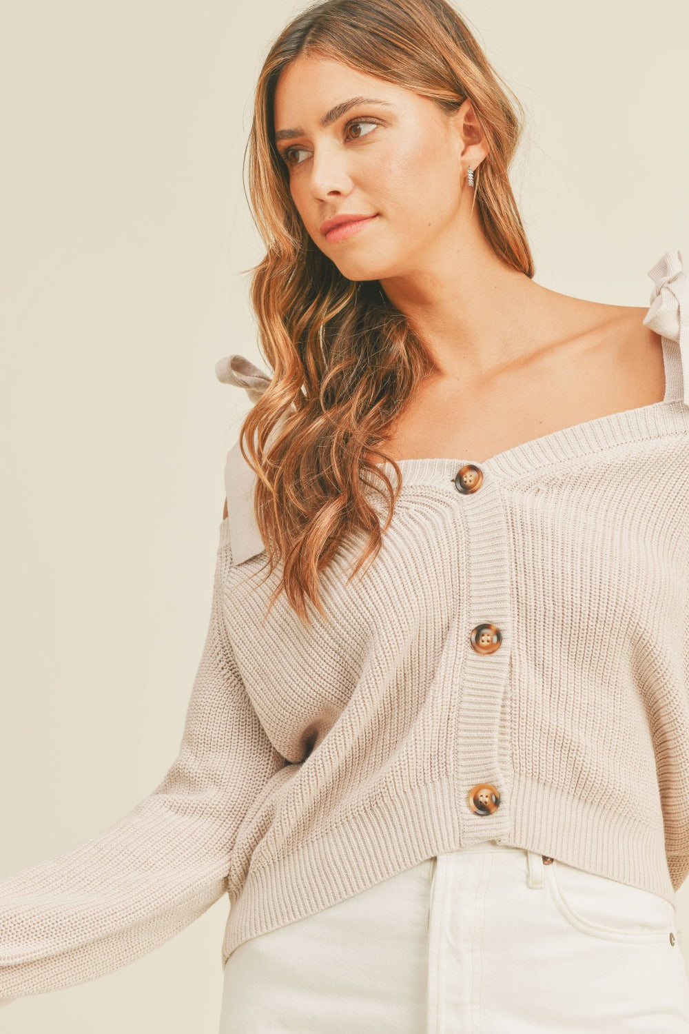 A woman with long, wavy hair is wearing a MABLE Off Shoulder Self Tie Strap Button Down Sweater in beige, paired with white pants, standing against a plain background.