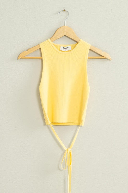 The Good Times Ribbed Sweater Tank Top, featuring a light green color, cropped hem, and back tie, hangs on a wooden hanger against a beige wall.