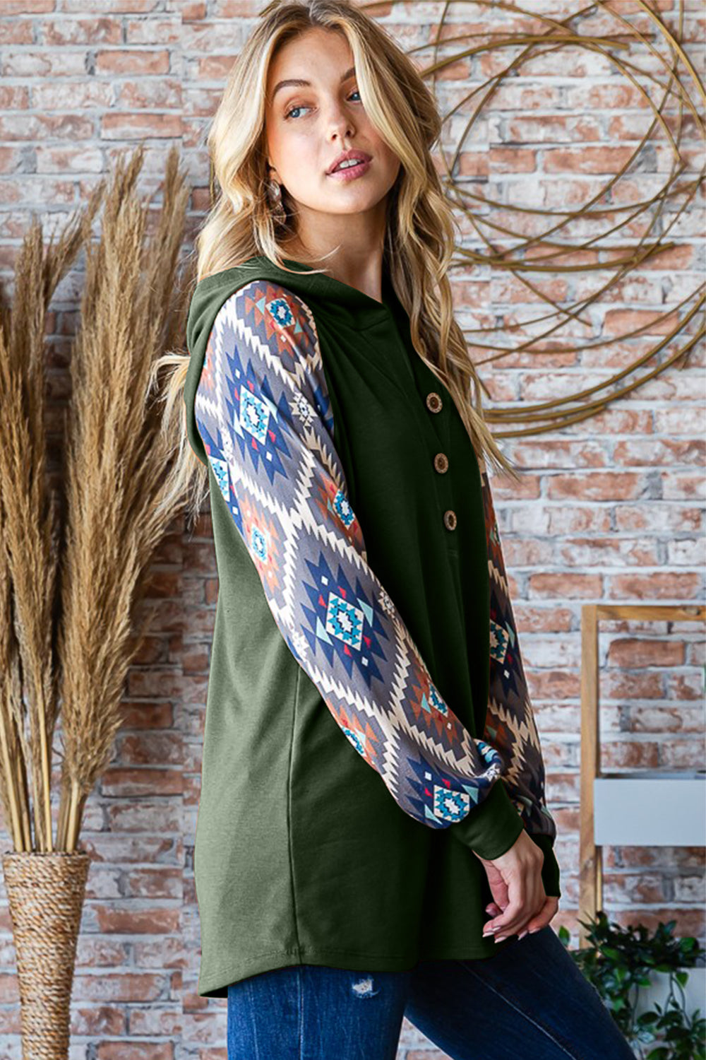 A woman poses indoors wearing a Heimish Full Size Half Button Printed Long Sleeve Hooded Top, showcasing its unique sleeve print design. Holding the hood with one hand, she complements her casual and on-trend outfit with blue jeans. The background features brick walls and tall dried plants.