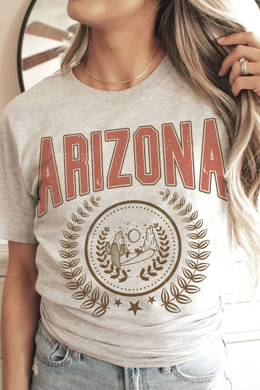 A person wearing a light gray ARIZONA Graphic Tee that showcases a desert scene with cacti and a sun, encircled by a laurel wreath design. This tee is made from 100% cotton and is available in unisex sizing.