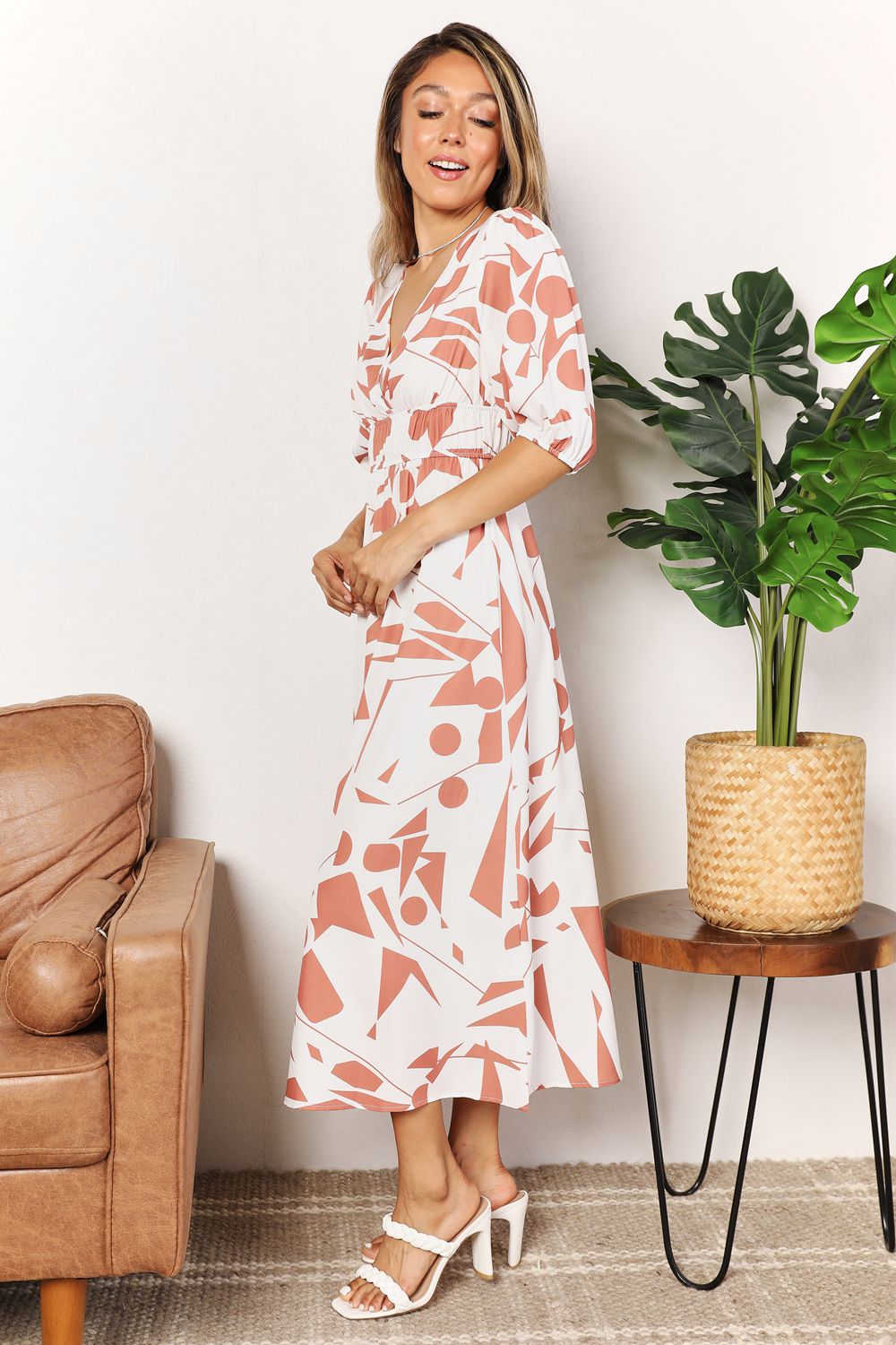 A person in a Printed Surplice Balloon Sleeve Dress stands near a brown chair and a round table with a potted plant.