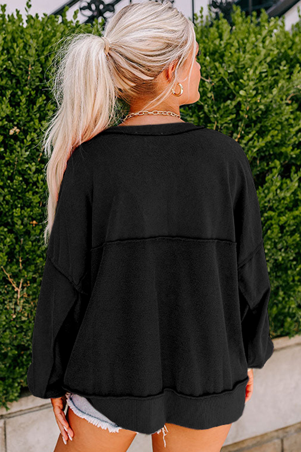 A person with long blonde hair in a ponytail, wearing a black Drop Shoulder Henley Buttons Sweatshirt and shorts, stands outdoors with greenery in the background.