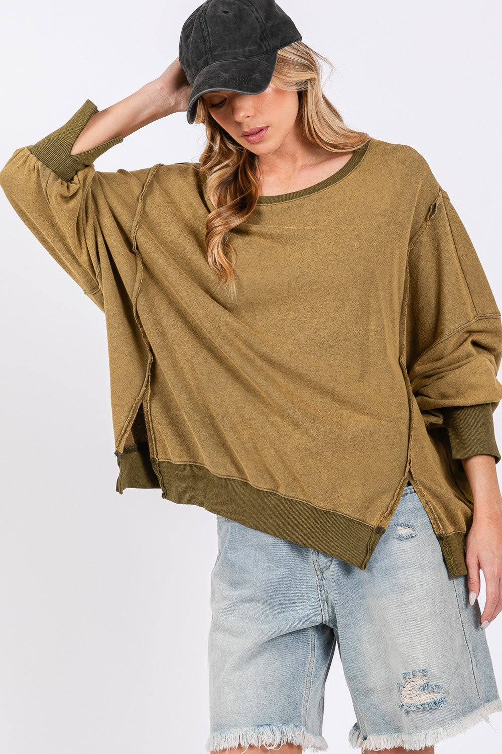 A person wearing a loose, oversized mineral wash SAGE + FIG Side Slit Sweatshirt in olive green, light blue jeans, and a black cap poses against a plain background.