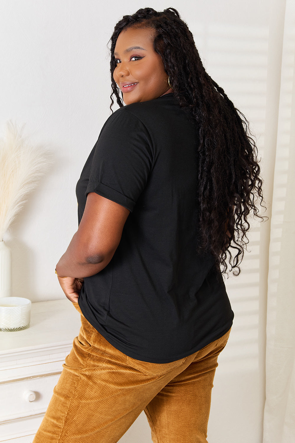 A woman stands with her hands in the pockets of her brown corduroy pants, wearing a casual Simply Love BE KIND Graphic Round Neck T-Shirt that reads "BE KIND" in large colorful letters, showcasing positive message clothing.