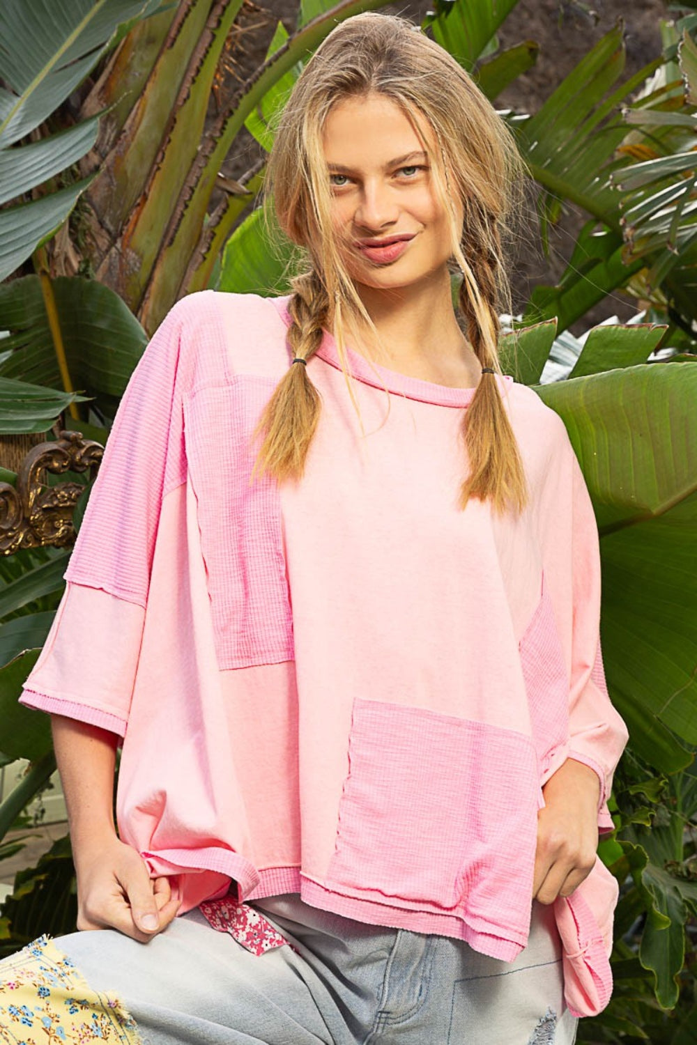 A person with braided hair is wearing a POL Frayed Edge Patchwork Oversized T-Shirt in pink, paired with loose jeans. They stand outside, surrounded by lush green foliage.