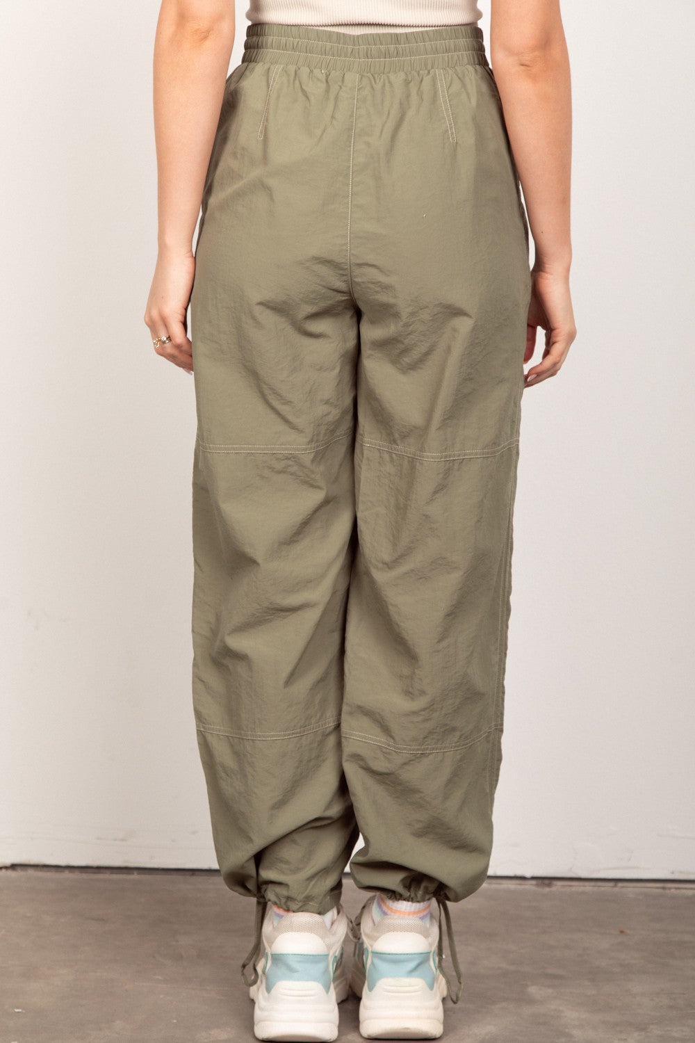 A person wearing VERY J Drawstring Woven Parachute Joggers in olive green and white sneakers stands against a plain background, showcasing the seamless blend of style and comfort with intricate woven fabric details.