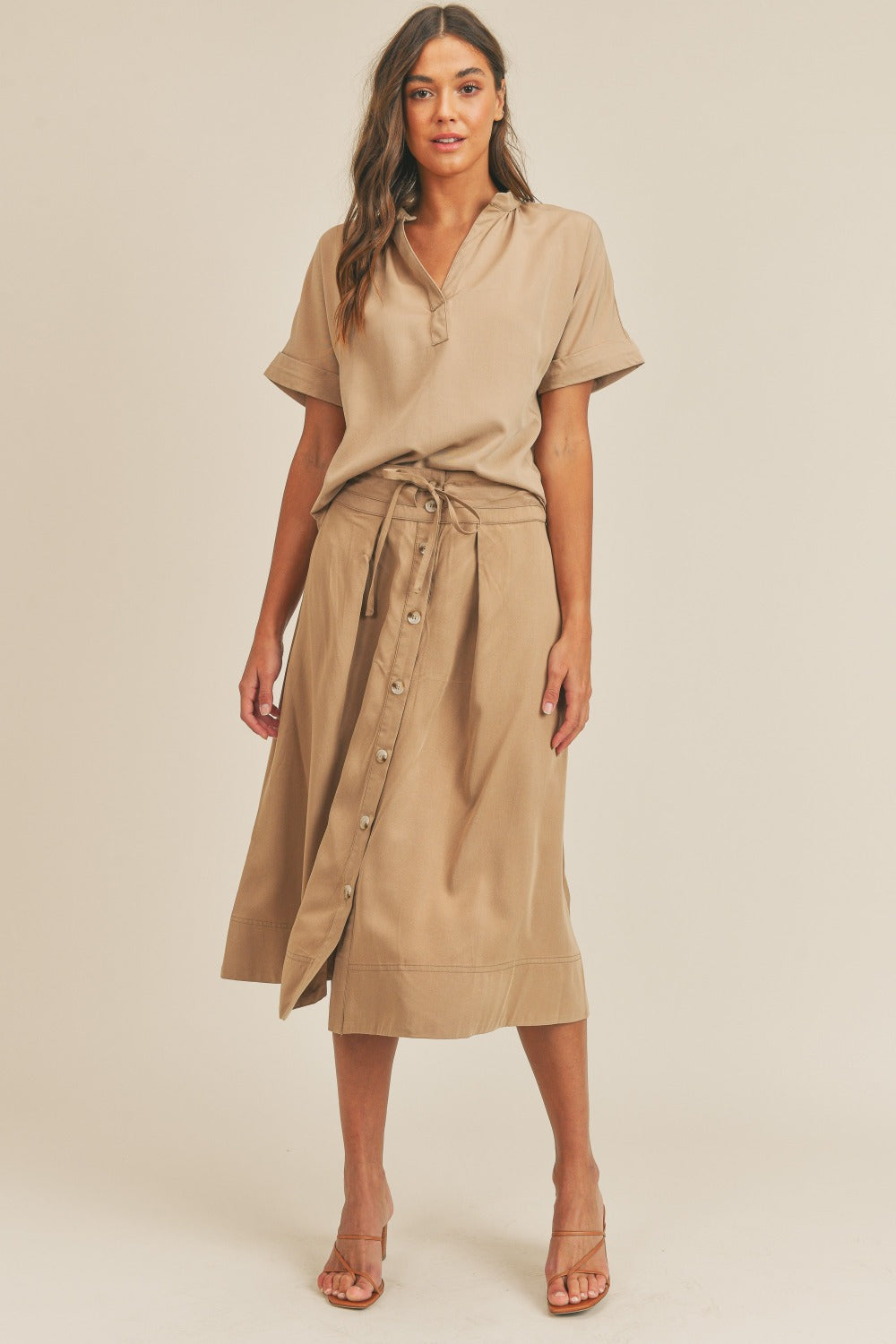 A woman is standing against a plain background wearing the MABLE Short Sleeve Top and Button Down Midi Skirt Set in beige. The elegant two-piece set, which includes a belted midi skirt, is paired perfectly with beige heels as she confidently poses with one hand on her hip.