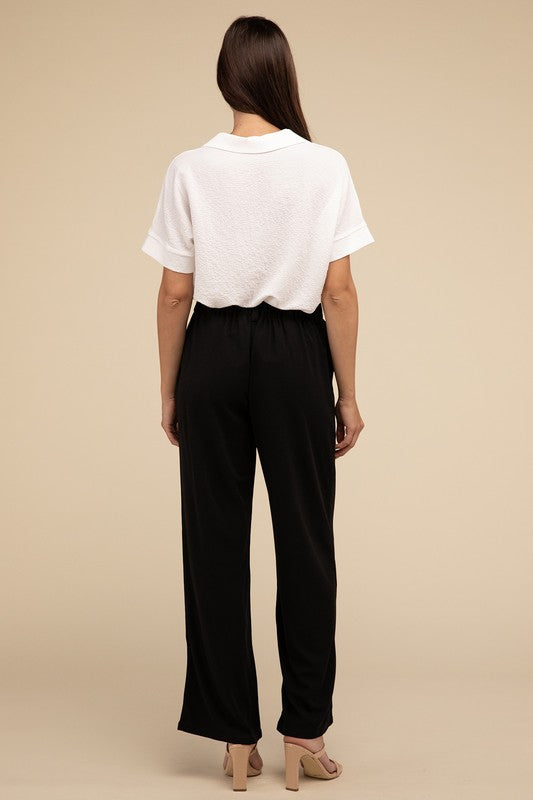 A woman stands against a beige background, highlighting her chic ensemble with a tailored fit. She wears a white short-sleeve shirt paired with versatile pink Waffle Trouser Pants, hands casually in pockets, complemented by stylish strappy sandals.