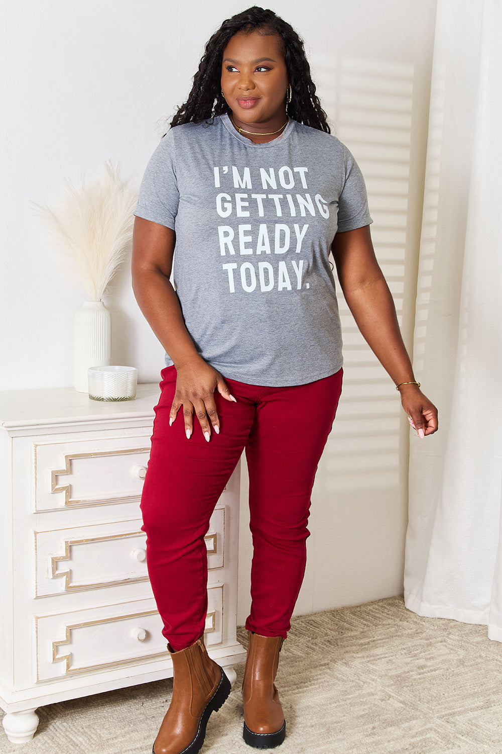 Dressed in a Simply Love I'M NOT GETTING READY TODAY Graphic T-Shirt and vibrant red pants, the individual exudes a carefree style while standing near a white surface adorned with decorative objects.