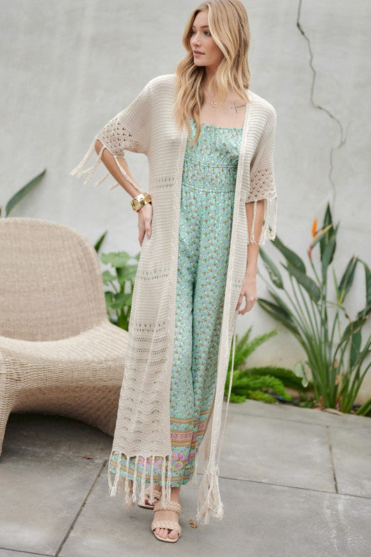 A woman in a green floral dress and a Solid Long Cardigan With Fringe exudes boho-chic vibes as she strolls on a patio, surrounded by outdoor furniture and lush plants.