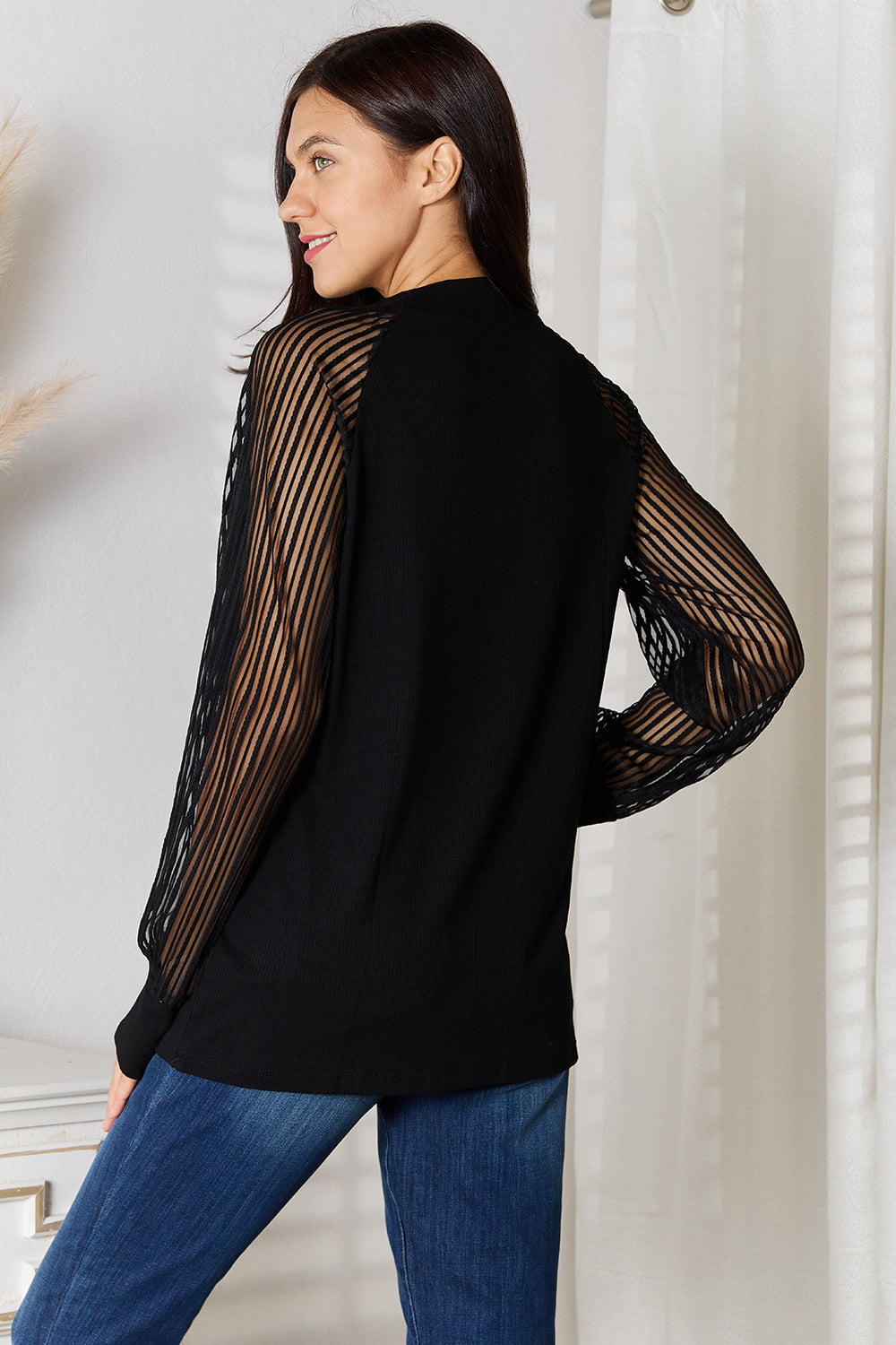 A woman with long dark hair stands indoors wearing a versatile wardrobe piece: the Double Take Round Neck Raglan Sleeve Blouse paired with blue jeans, her right hand in her pocket. A white vase with pampas grass is on the table next to her.