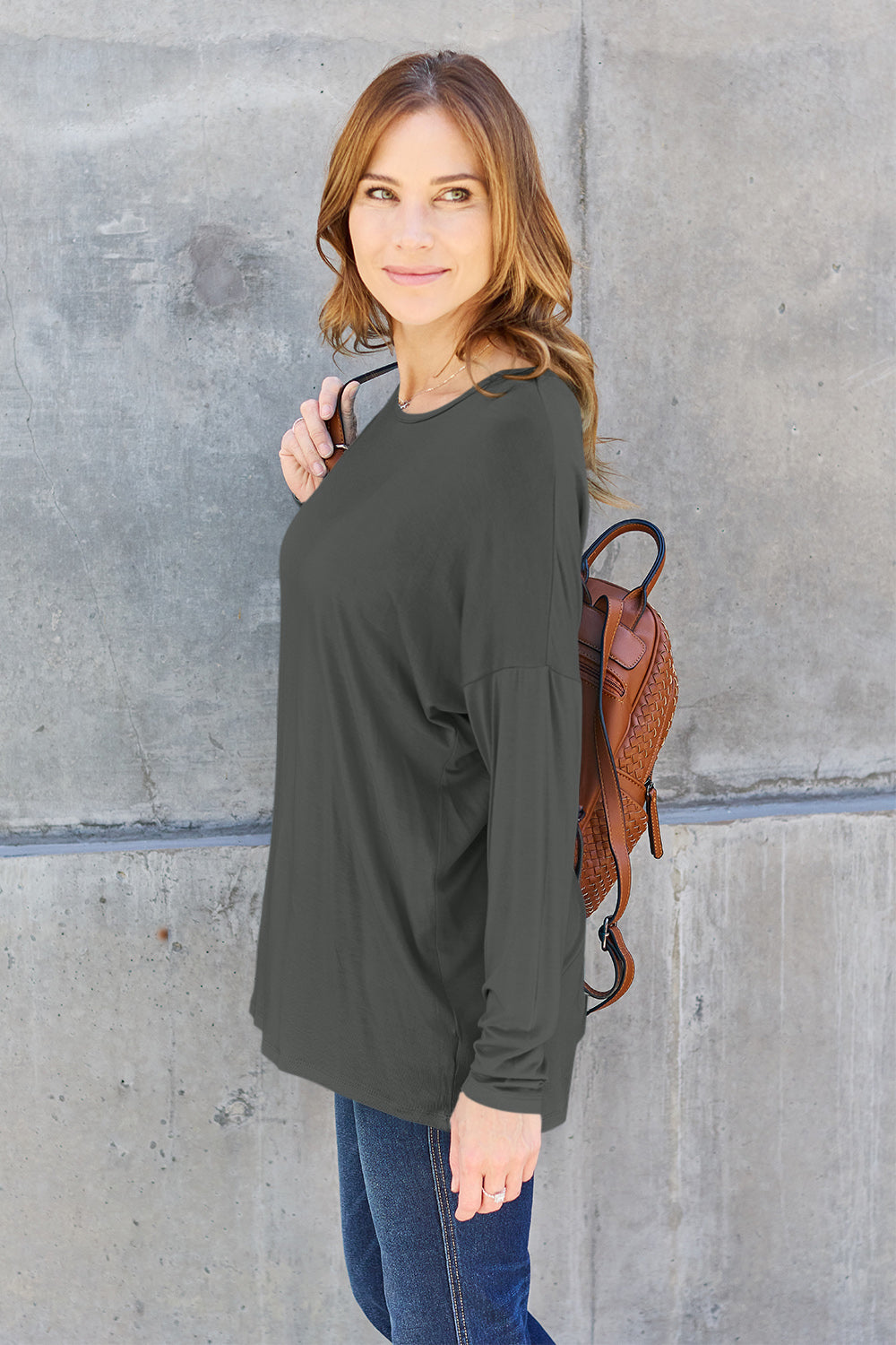 A woman with shoulder-length hair wearing a Basic Bae Full Size Round Neck Dropped Shoulder T-Shirt in green and a pair of jeans, exuding a basic style, stands against a concrete wall. She's holding an imported brown woven backpack on her shoulder while touching her hair with her hand.