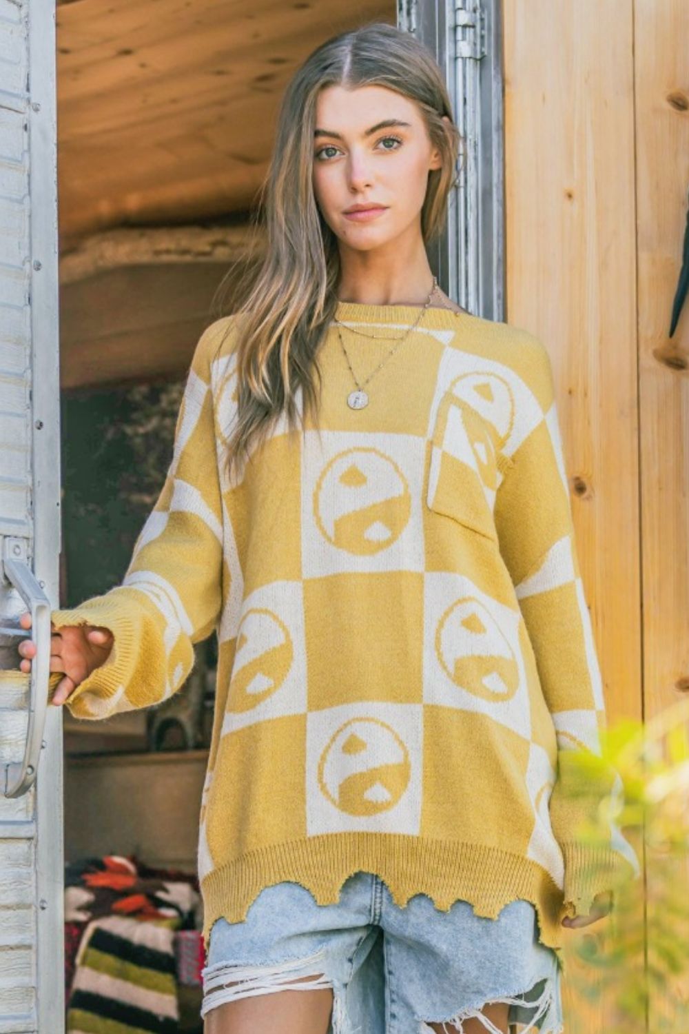 A woman wearing the And The Why Checkerboard Sweater with a Yin Yang pattern and distressed denim shorts stands in a doorway.