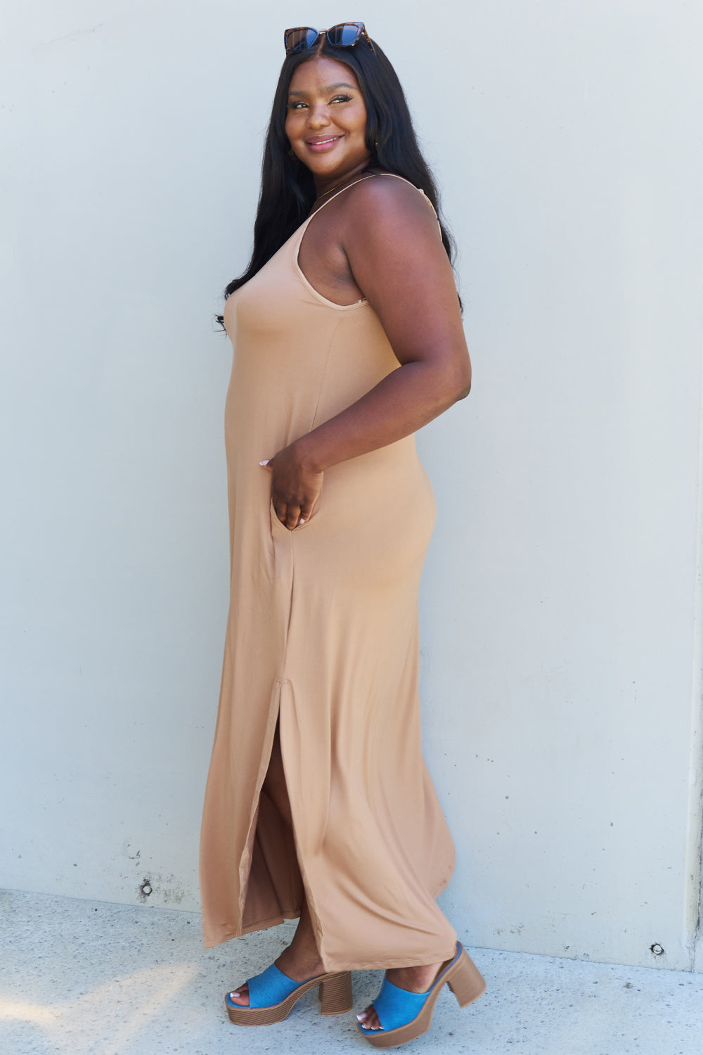 A woman stands against a plain background, wearing the Ninexis Good Energy Full Size Cami Side Slit Maxi Dress in Camel along with a wide-brimmed hat. She has one hand on her hip and is smiling.