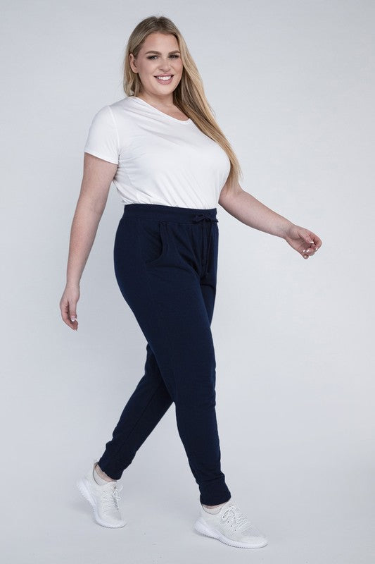 A person stands against a white background, wearing a white t-shirt, grey Plus-Size Jogger Pants with an adjustable waistband, and white sneakers. They have long blonde hair and are posing with one hand on their hip.