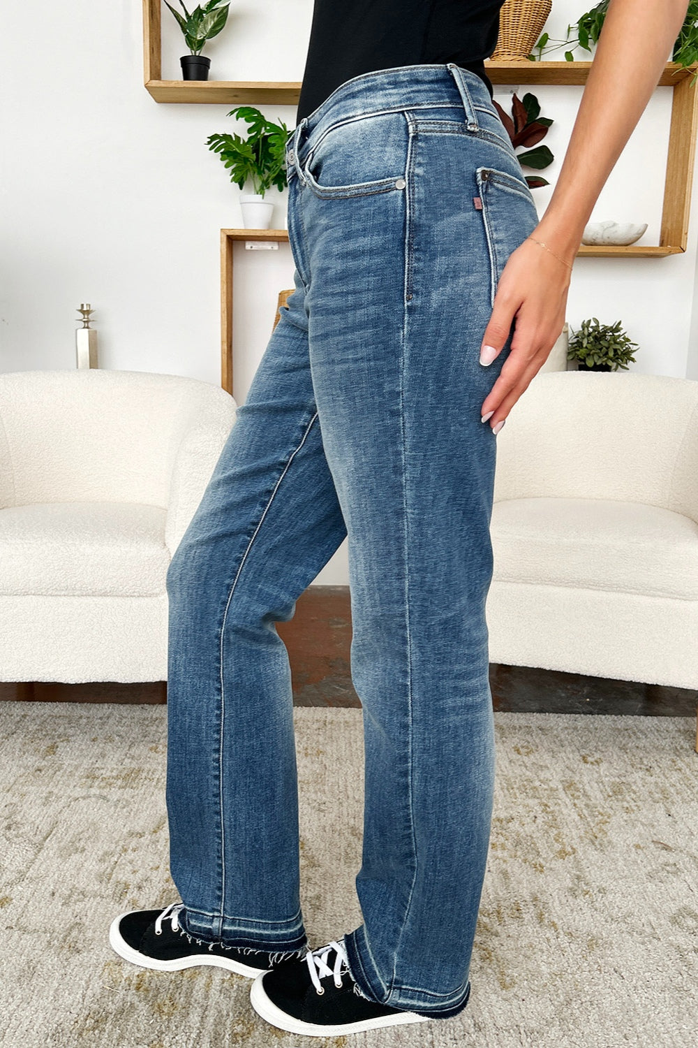 A person standing indoors, wearing a pair of Judy Blue Full Size Mid Rise Release Hem Jeans and black sneakers, holding the loose fabric of the jeans on one side.