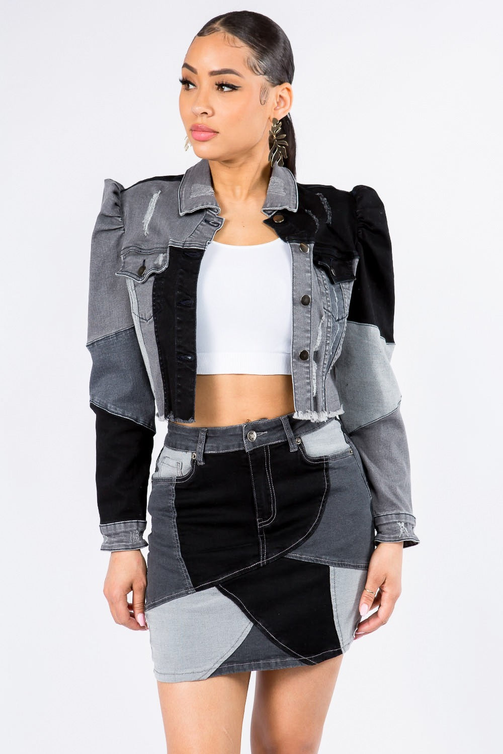 A woman is wearing the American Bazi Button Up Cropped Patchwork Denim Jacket, featuring puff shoulders and a raw hem, paired with a matching skirt in black, grey, and white. Underneath, she sports a white crop top while standing against a plain white background.