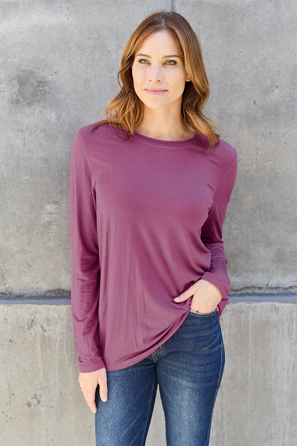 A woman with shoulder-length hair wearing the Basic Bae Full Size Round Neck Long Sleeve Top and blue jeans stands in front of a concrete wall, holding a beige handbag over her shoulder. Her outfit exemplifies basic style, with the top made from 100% polyester and recommended for machine wash cold.