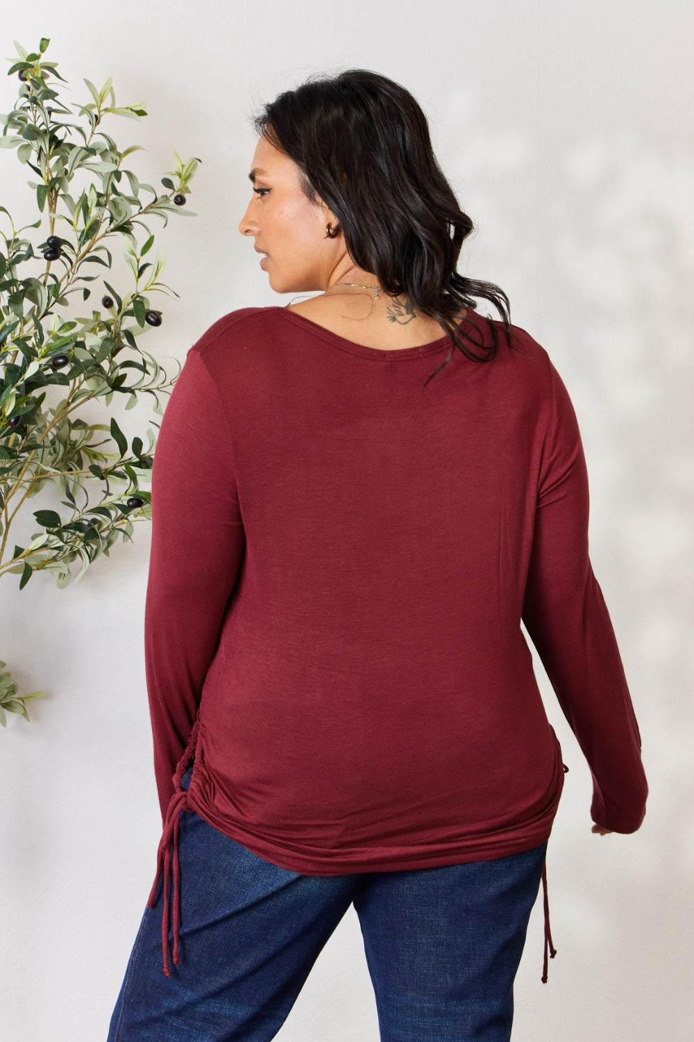 A woman stands in front of a plain background and a small tree, wearing the Culture Code Full Size Drawstring Round Neck Long Sleeve Top in maroon and blue jeans.