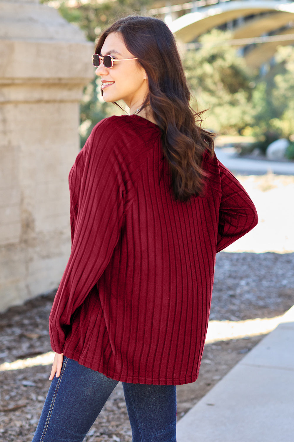 A woman with long brown hair wearing sunglasses and a Basic Bae full-size, slightly stretchy ribbed round neck long sleeve knit top in red, paired with blue jeans, is standing outdoors.