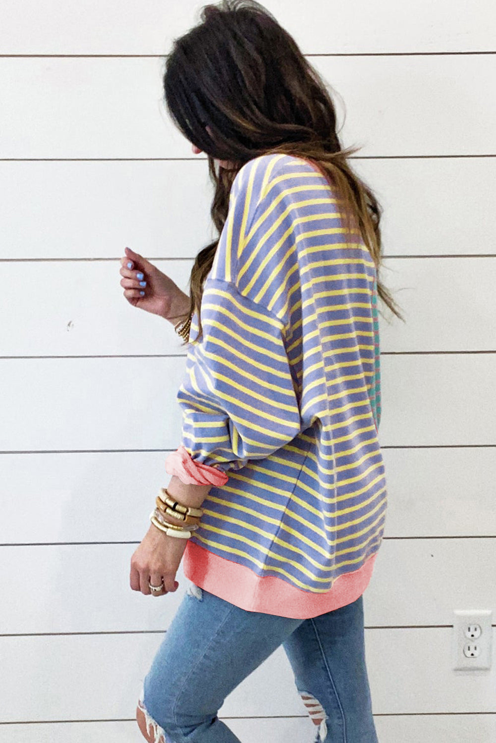 A person with wavy hair is seen from behind wearing a Casual Stripe Colorblock Drop Shoulder Oversize Sweatshirt in pastel colors and blue jeans. They are carrying a white shoulder bag, showcasing a trendy color block design.