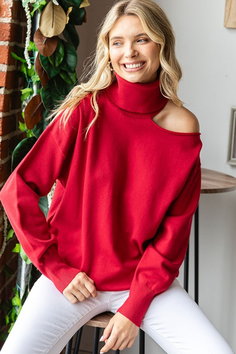 A blonde woman in the stylish First Love Cut Out Shoulder Turtleneck Sweater and white pants sits on a stool against a wall, with plants in the background.