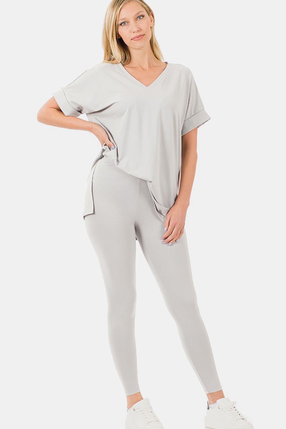 The individual gracefully dons the Zenana V-Neck Rolled Short Sleeve T-Shirt and Leggings Lounge Set in white, standing confidently with one hand on their hip. Paired with sandals, this ensemble captures both comfort and style against a simple backdrop.