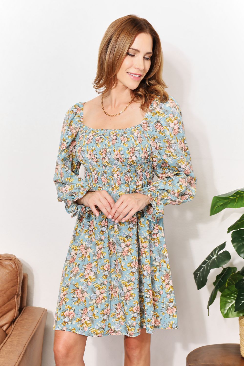 A woman in the Honey Floral Smocked Flounce Sleeve Square Neck Dress stands indoors, holding the hem of her dress. She is smiling slightly with her eyes looking downward. Nearby, a plant and a brown couch complete the cozy scene.