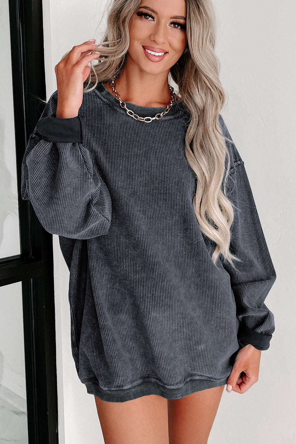 A person with long, blonde hair smiles while wearing an oversized Gray Solid Ribbed Knit Round Neck Pullover Sweatshirt and a gold chain necklace. They are standing indoors near a window, embodying versatile style.