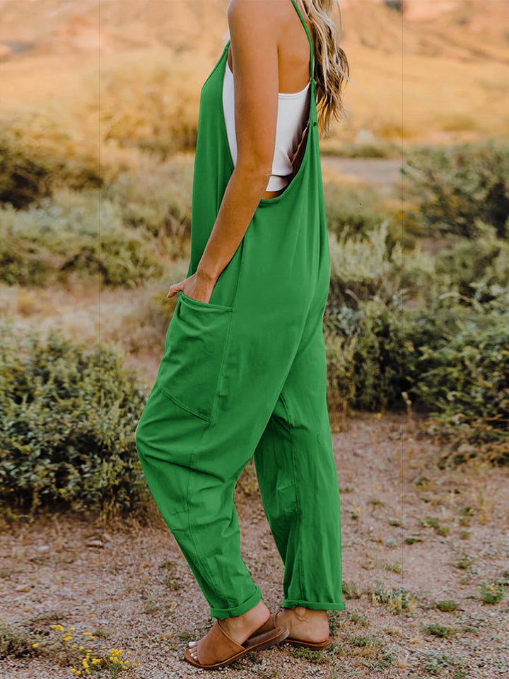 A woman wearing the Double Take Full Size Sleeveless V-Neck Pocketed Jumpsuit in green and a beige cap stands outdoors in a desert landscape with plants and rocky cliffs in the background.