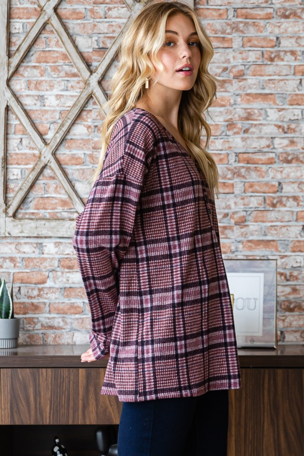 Sporting long blonde hair, a person stands against a brick wall wearing the Heimish Full Size Plaid V-Neck Long Sleeve Top in pink and black. Framed butterfly prints adorn the background, while this adaptable wardrobe item adds a touch of flair to any ensemble.